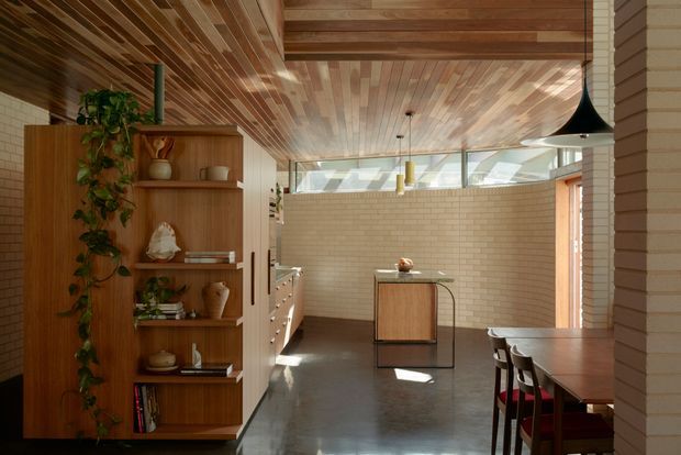 Warm, open-plan interior with a timber-lined ceiling and matching timber built-in shelving, a compact kitchen or dining zone, exposed cream brick walls, polished dark concrete flooring, and clerestory glazing. A long dining table with stools occupies the right foreground, while a small island or console sits centrally. The space has strong mid-century-inspired detailing, natural materials, and abundant filtered daylight.