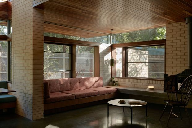 A warm mid-century-inspired living room with a built-in tufted sofa beneath a timber-lined ceiling. Exposed cream brick piers and walls frame expansive dark timber windows, while a dark polished floor, round coffee tables, and a black lounge chair create a restrained, earthy interior. Abundant glazing brings in filtered natural light and views of surrounding trees.