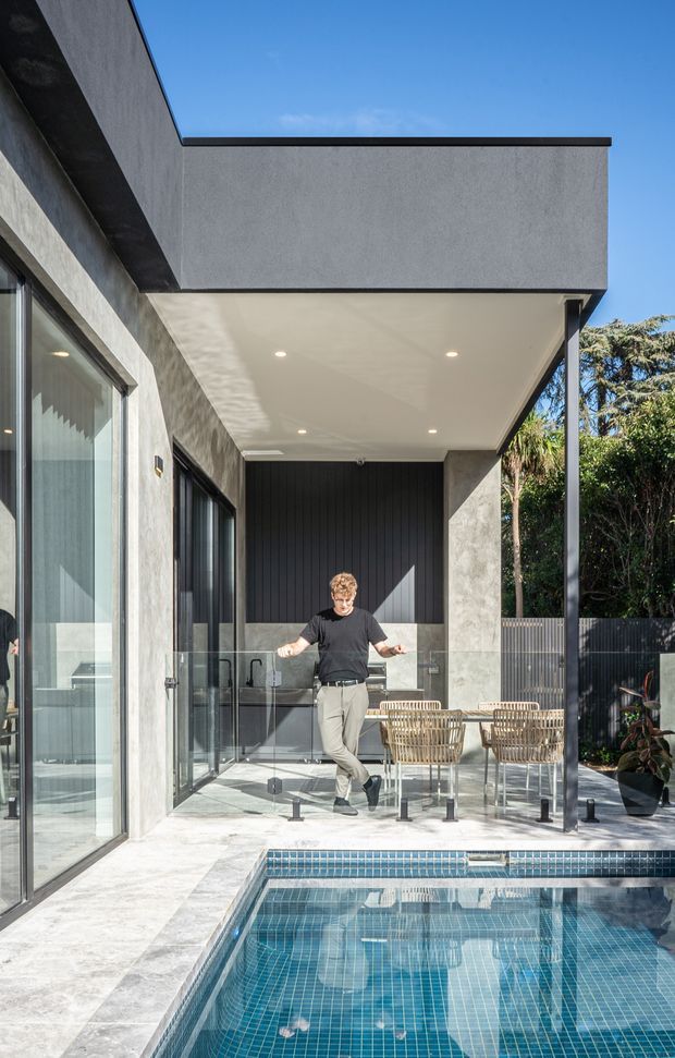 A contemporary outdoor entertaining area beside a tiled swimming pool, enclosed by a flat-roofed concrete-rendered home. The covered terrace features a pale tiled floor, recessed ceiling lights, black vertical wall cladding, extensive dark-framed glazing, a glass pool barrier, and woven outdoor dining furniture. Strong sunlight, greenery, and the pool create a refined indoor-outdoor connection.