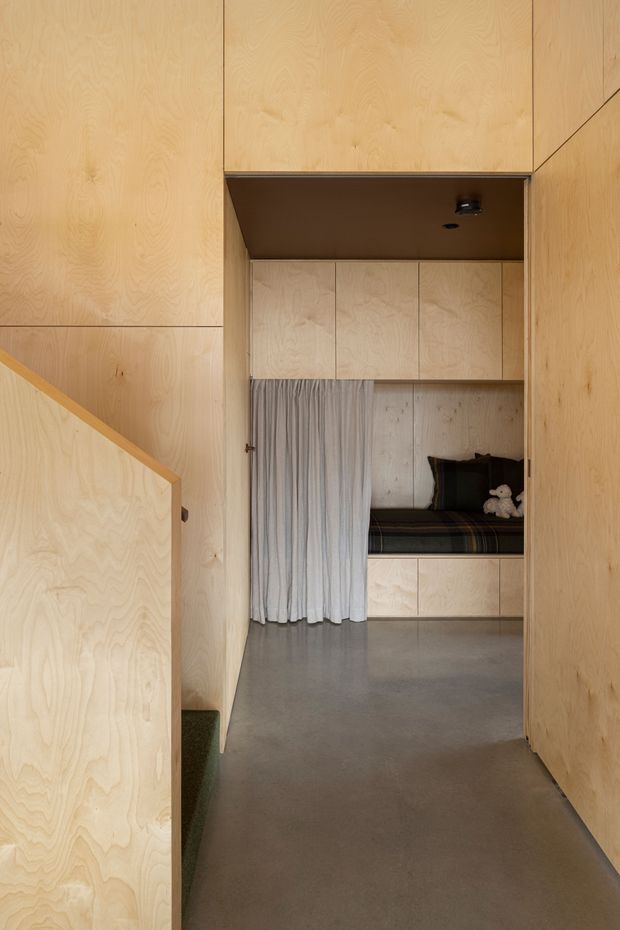 A compact, highly integrated interior viewed from a plywood-lined hallway or stair landing. Pale timber plywood panels cover the walls, stair balustrade, ceiling bulkhead, doors, and built-in joinery. Ahead is a small sleeping nook or bedroom with overhead cabinets, a built-in bed, curtain divider, dark cushions, and a small stuffed toy. The floor is a smooth grey concrete-like finish, and the overall design is minimalist and space-efficient.