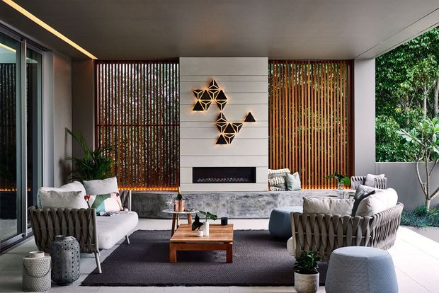 A sophisticated covered outdoor lounge or indoor-outdoor living area with a large sectional seating arrangement, woven lounge chairs, a timber coffee table, and a linear fireplace set into a pale stone feature wall. Vertical timber screening, extensive glazing, integrated warm lighting, and lush greenery create a refined connection to the garden.