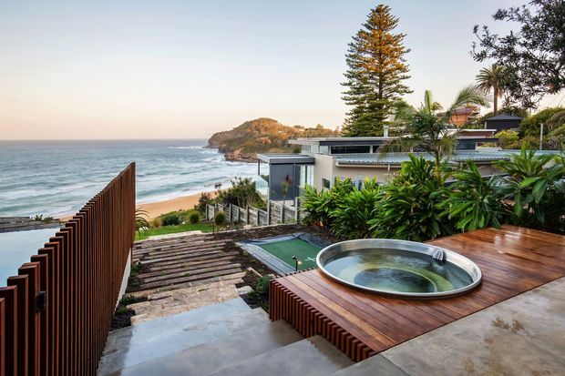 A contemporary coastal home viewed from an elevated outdoor terrace, overlooking a sandy beach, ocean and rocky headland. The foreground features a timber-decked rooftop or terrace with a circular built-in spa, concrete steps, vertical timber screening, lush tropical planting and a small pool or water feature. The house has flat rooflines, modern glazing and a layered indoor-outdoor design.