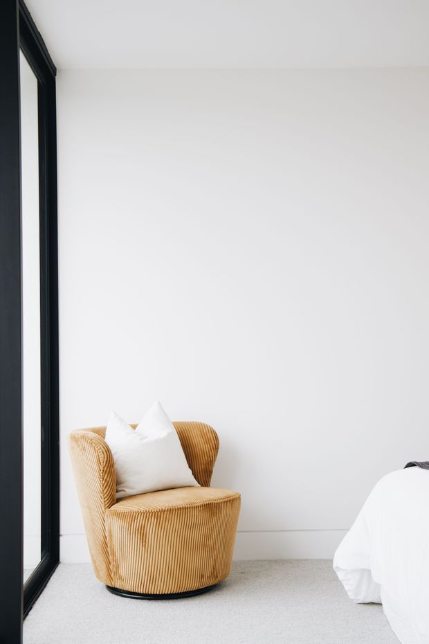 Minimal bedroom corner with a mustard-gold ribbed upholstered armchair beside a white bed, set against plain white walls and a light carpeted floor. A tall black-framed window or glazed door provides a strong vertical architectural element, creating a bright, uncluttered interior.