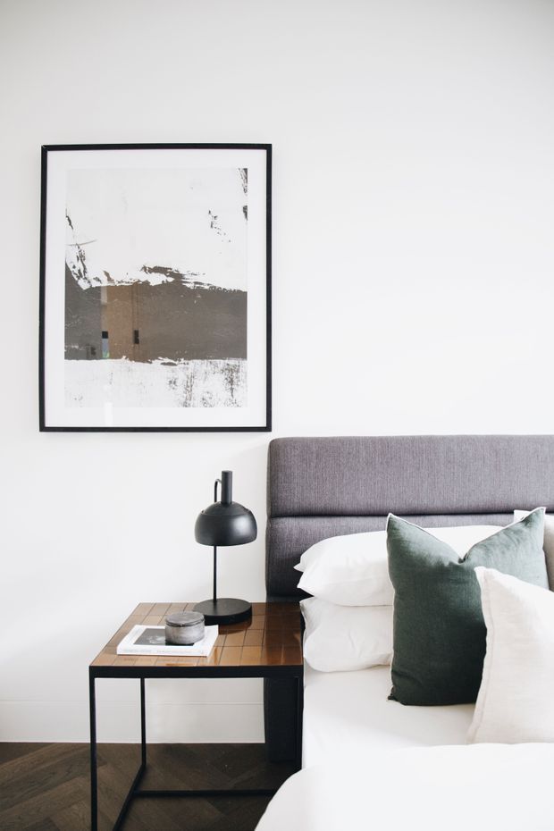 A refined modern bedroom vignette with a grey upholstered bed, crisp white bedding, and deep green accent cushions. A black metal bedside table with a wood-toned top supports a sculptural black lamp and small decor. The room has white walls, dark timber flooring, and a minimalist black-framed abstract artwork.