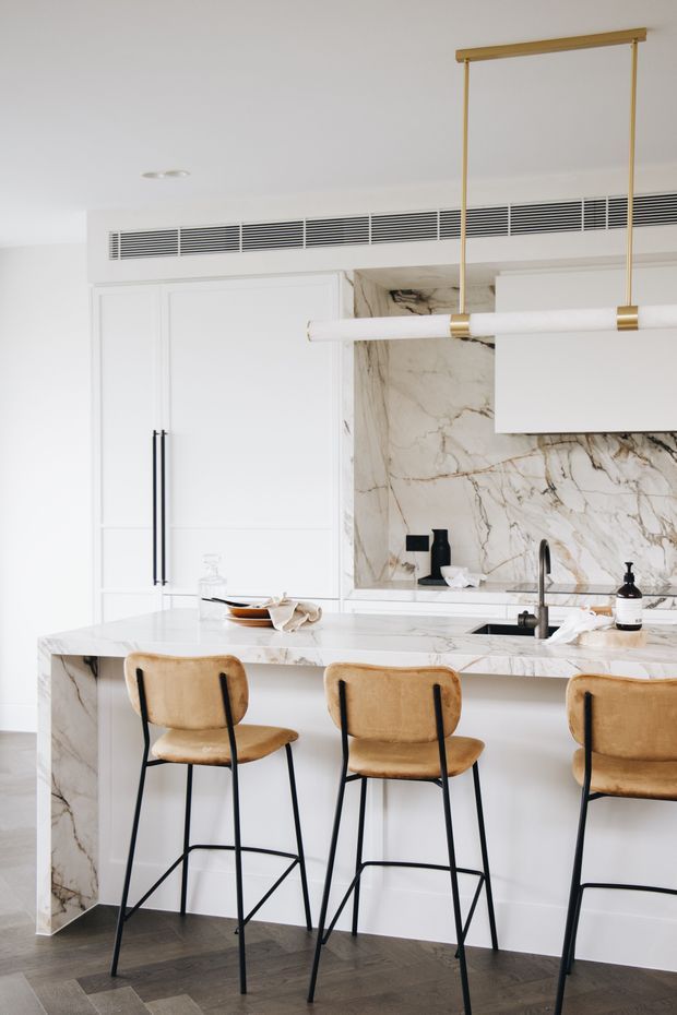 Bright contemporary kitchen with white paneled cabinetry, a large white marble island and matching marble splashback, black fixtures, and three tan upholstered bar stools. A linear white pendant light with brass suspension hangs above the island, while a concealed linear air-conditioning grille runs across the upper cabinetry. Dark herringbone timber flooring provides contrast.