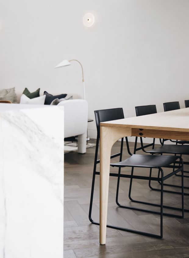 Bright, minimalist open-plan interior showing a pale timber dining table with black metal-framed chairs in the foreground, alongside a white sofa with neutral and dark cushions. The room has white walls, a slim floor lamp, light wood flooring, and a pale stone or marble kitchen island edge at left.