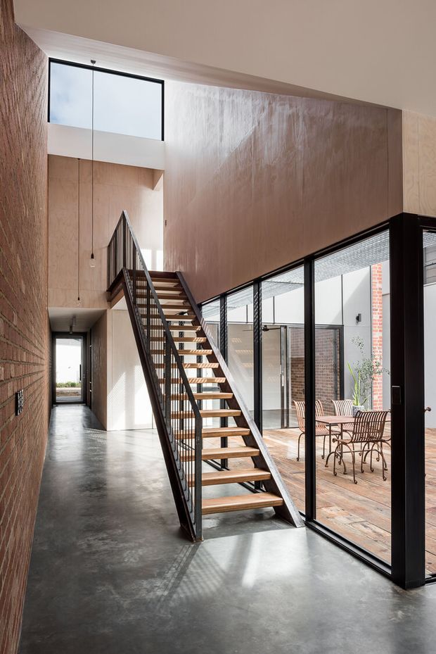 A tall, light-filled interior circulation space with an open-riser timber staircase and dark metal balustrade. The space combines polished concrete flooring, warm timber wall panelling, exposed brick, black-framed floor-to-ceiling glazing, and a visible brick-paved courtyard with outdoor furniture beyond. A high-level window and strong natural light emphasize the vertical volume.