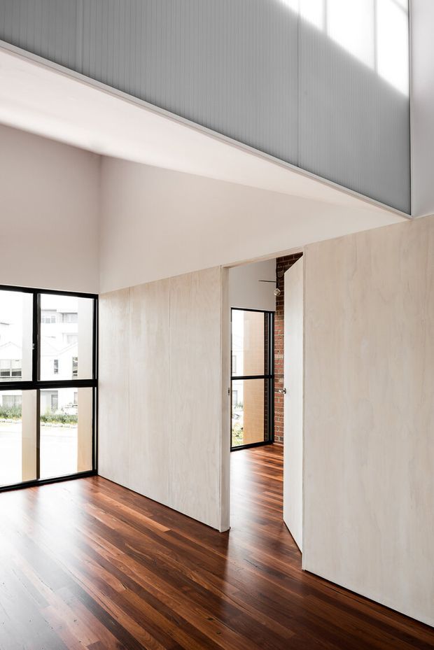 Bright, minimalist interior with an open-plan circulation area and tall black-framed glazing. Dark timber flooring contrasts with pale, smooth wall panels and a high ceiling featuring a grey vertically ribbed surface. A partial exposed brick wall is visible through the doorway, adding subtle texture. The image appears to show a hallway or landing adjoining another room.