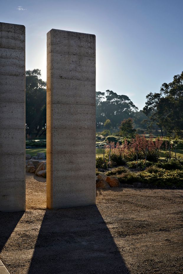 An exterior landscape photograph featuring tall, freestanding raw concrete walls or architectural panels arranged in a garden setting. Warm low-angle sunlight creates strong elongated shadows across the gravel or compacted-earth ground. Native-looking trees, shrubs, planted beds, and large rocks surround the sculptural concrete elements, giving the scene a quiet, monumental, garden-installation character.