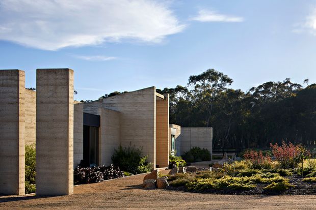 Exterior architectural view of a minimalist, monolithic concrete residence or building set within a landscaped site. Tall vertical concrete walls and projecting planes create a sculptural facade, with dark recessed glazing and a low, horizontal concrete volume extending into the background. Gravel paving, rocks, low shrubs, and ornamental planting surround the structure, with mature woodland beyond under a clear sky.
