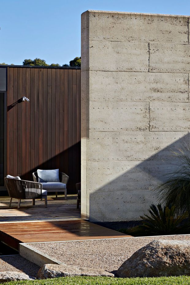 A modern residential courtyard or outdoor living area featuring a tall board-formed concrete wall, dark vertical timber cladding, and a timber deck. Two woven outdoor lounge chairs with cushions sit beside the building, while gravel landscaping, large rocks, and sculptural grasses frame the foreground. Strong sunlight creates angular shadows across the concrete wall and deck.