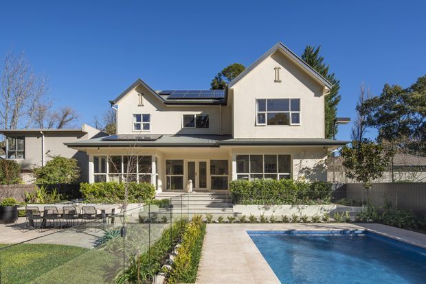 A two-storey residential home with a cream rendered facade, pitched gable roofs, solar panels, and a covered rear veranda. The landscaped backyard includes a paved entertaining area, glass pool fencing, and a rectangular swimming pool.
