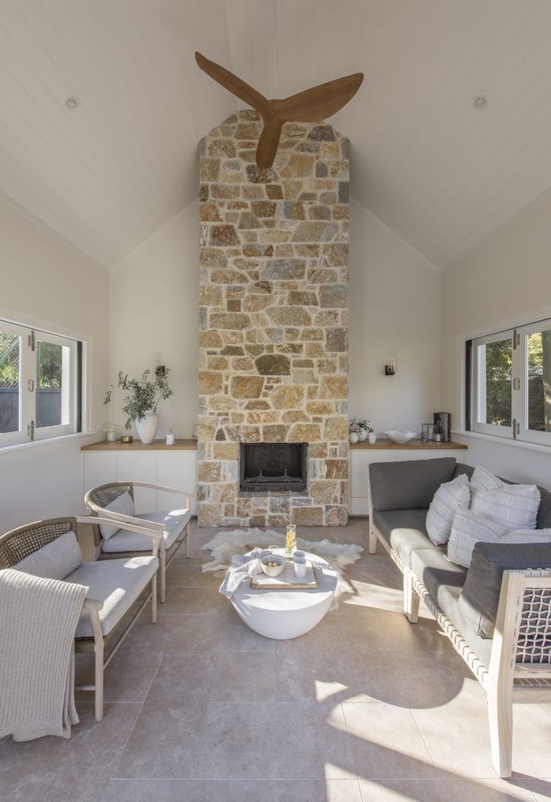 Bright, airy living room centered on a tall exposed-stone fireplace with a dark timber mantel shelf and sculptural timber feature above. The room has a vaulted white panelled ceiling, pale tiled flooring, white walls, large windows, and light woven lounge furniture arranged around a small white coffee table.