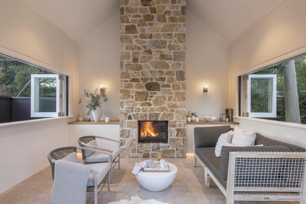 A bright, narrow sunroom or enclosed outdoor lounge with a vaulted white ceiling, large opening windows, and a prominent full-height natural stone fireplace. The room is furnished with light woven lounge chairs, a grey sofa, a low white coffee table, and subtle greenery, creating a relaxed indoor-outdoor atmosphere.