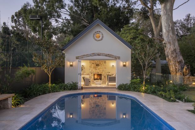 A small gabled pool house or cabana opens directly onto a swimming pool. The white rendered facade has dark trim, a circular feature window, wall sconces, and a broad glazed opening with folding doors. Inside, a warm stone feature wall and fireplace are visible. The pool is surrounded by pale paving and landscaped gardens with mature trees and screening fencing.