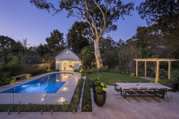 Twilight view of a landscaped residential backyard with a long rectangular swimming pool, paved pool terrace, outdoor dining area beneath a timber pergola, and a small illuminated gabled garden pavilion. Mature trees and planting frame the garden.