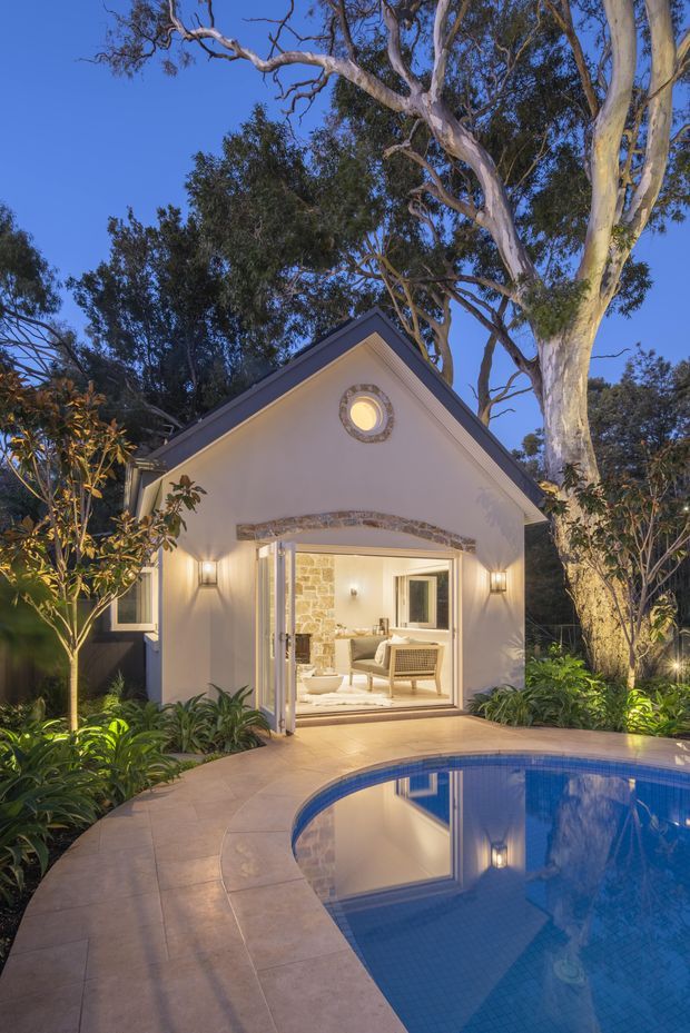 A dusk exterior view of a small, gable-roofed garden pavilion or poolside guesthouse surrounded by mature eucalyptus trees and lush planting. The light-coloured rendered facade has a circular feature window, stone-trimmed arched opening, wall lights, and fully open glazed doors revealing a warmly lit seating area inside. A curved tiled pool occupies the foreground with a paved terrace around it.
