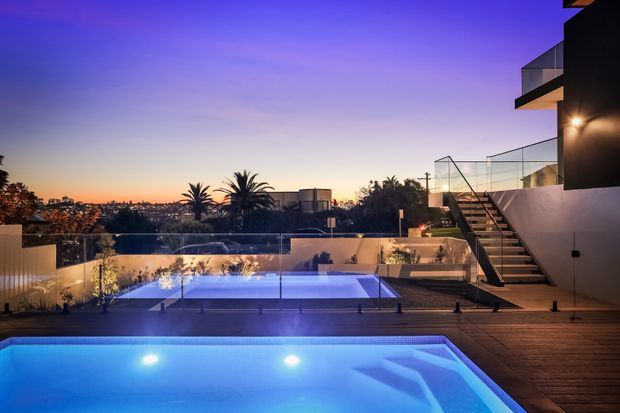 Twilight exterior view of a contemporary multi-level residence with a brightly illuminated swimming pool in the foreground, timber decking, glass balustrades, landscaped planting, and an elevated terrace with external stairs. The home overlooks a suburban cityscape and is presented in a dramatic blue-purple evening light.