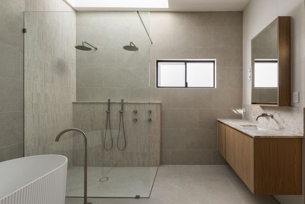 A contemporary bathroom with a walk-in double shower enclosed by clear glass, a freestanding white bathtub in the foreground, and a timber-veneered floating vanity with a pale stone benchtop. Large-format beige-grey wall and floor tiles, a skylight, and a small horizontal window create a bright, refined space. A wall-mounted mirrored cabinet is visible above the vanity.