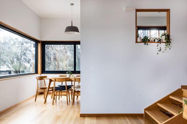 Bright contemporary interior showing a compact dining area beside a stairway. The dining space has pale timber flooring, white walls, large black-framed windows, a timber dining table with matching chairs, and a simple dark pendant light. At right, timber stairs rise beside a white wall with a timber-framed interior window and trailing plant.