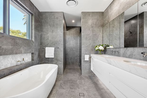 Contemporary bathroom featuring large-format grey stone-look wall and floor tiles, a freestanding white bathtub, frameless glass shower, double vanity with a white marble-look benchtop, large wall mirror, and recessed ceiling lighting. Natural light enters through the window, creating a bright, refined space.
