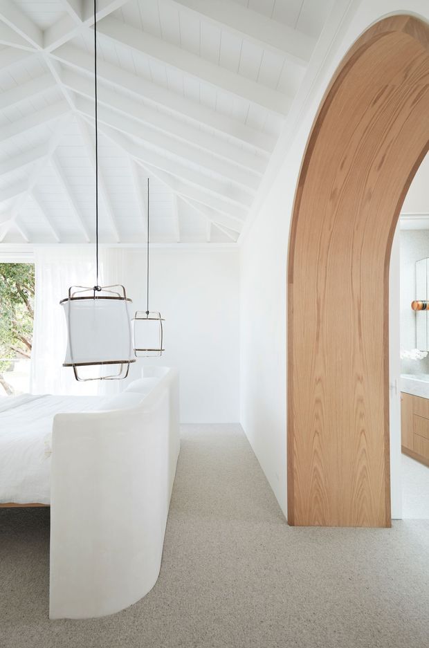 Bright, predominantly white bedroom with a dramatic vaulted ceiling featuring exposed painted timber rafters and panelled lining. A curved white bed surround or partition occupies the foreground, with minimalist suspended pendant lights. A large arched timber doorway frames a glimpse into an adjoining bathroom with timber cabinetry and a pale stone or tiled benchtop.