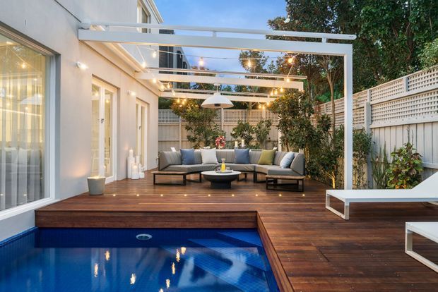 A contemporary outdoor entertaining area beside a modern white-rendered house, featuring a timber deck, rectangular swimming pool, built-in outdoor lounge seating, white pergola, pendant string lighting, privacy screening, and lush planting. The space has a refined resort-like feel and is photographed at dusk.