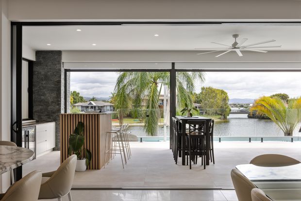 Contemporary open-plan living and dining area with floor-to-ceiling glazing overlooking a waterfront setting. The bright interior features a large dining table, bar-height seating, a marble-look kitchen island at left, dark textured wall cladding, light tiled flooring, and a covered outdoor terrace with a pool or water feature beyond.