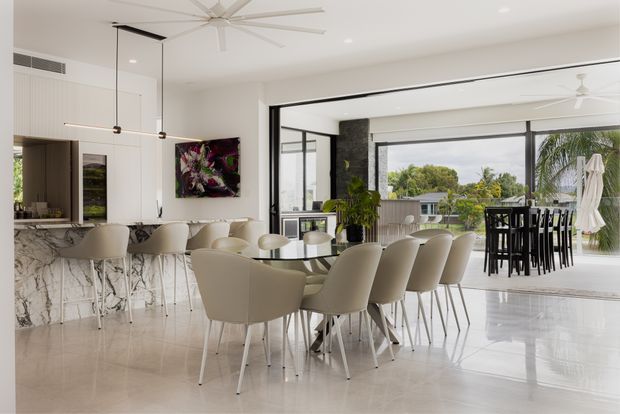 Bright, open-plan contemporary kitchen and dining area with a large marble-look island, curved upholstered bar stools, a long dining table with cream chairs, and expansive black-framed glazing opening to an outdoor terrace. White finishes, polished flooring, pendant lighting and ceiling fans create an airy, refined interior.