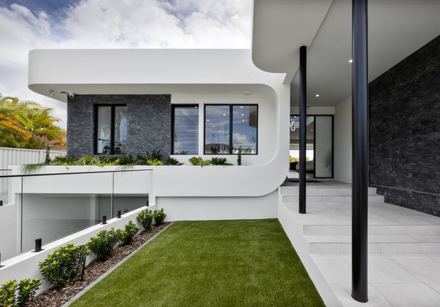 Contemporary multi-level residential exterior with sculptural white curved forms, extensive glazing, dark textured stone feature walls, slim black columns, and pale tiled terraces surrounding a manicured lawn and planted garden.