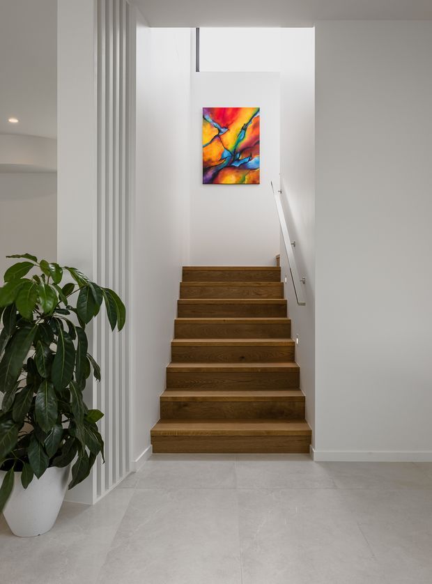 A bright, minimalist interior stairway with warm timber stairs ascending between white walls. A slim metal handrail lines the right side, while vertical white slatted screening defines the left edge. Large pale floor tiles and a potted leafy plant sit in the foreground, and a vivid abstract artwork provides a colourful focal point on the upper wall.
