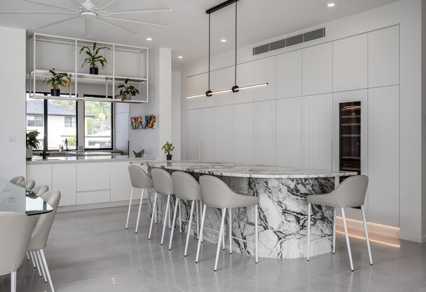 Bright contemporary open-plan kitchen and dining area with a large white marble-look island, integrated white cabinetry, bar seating, pendant lighting, indoor plants, and a dining table partially visible at left. The space has polished grey flooring, extensive built-in storage, and a large ceiling fan.