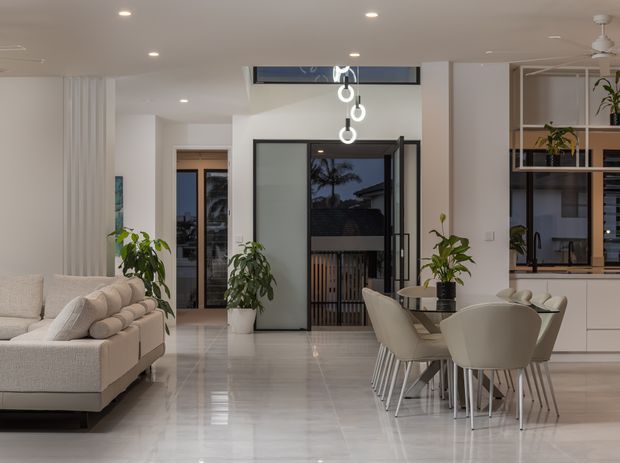 A bright, contemporary open-plan living and dining area with a neutral palette. The room features a large upholstered sectional sofa, a dining table with sculptural upholstered chairs, glossy light flooring, white walls and cabinetry, indoor plants, recessed ceiling lights, and a linear hanging pendant. Black-framed glazed doors open toward an outdoor area, while built-in shelving and cabinetry line the right side.