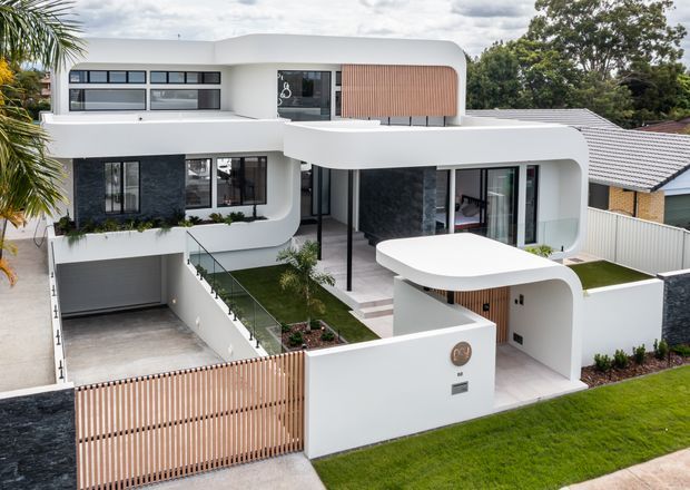 Contemporary multi-level suburban residence with a sculptural white rendered facade, rounded roof and balcony forms, extensive dark-framed glazing, charcoal stone feature walls, and warm vertical timber screening. The image shows a landscaped front yard, covered entry, driveway, and integrated garage.