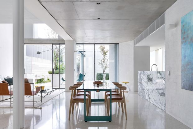 Bright open-plan contemporary interior showing a dining area in the foreground, with a glass-topped dining table and timber chairs. A living room is visible to the left, while full-height glazing opens toward a planted courtyard. The space features polished light stone or concrete flooring, white walls, a smooth exposed concrete ceiling, extensive glass, and a marble-clad kitchen island or counter. The restrained palette and abundant daylight create an airy, refined atmosphere.