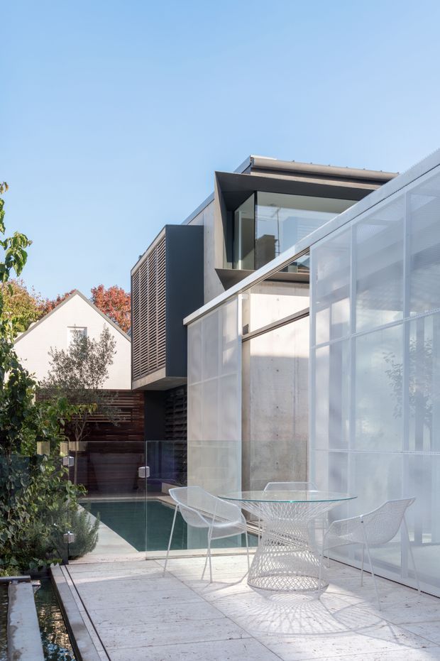 A modern residential courtyard or terrace viewed outdoors, featuring a pale paved deck with a glass dining table and white wire-frame chairs. The space is enclosed by translucent screened panels, clear glass balustrades, dark timber and metal architectural elements, and a concrete wall. A narrow linear water feature or pool edge runs beside the deck, with a neighbouring gabled house and planting visible in the background. The architecture has crisp geometric forms, extensive glazing, and strong indoor-outdoor connection.