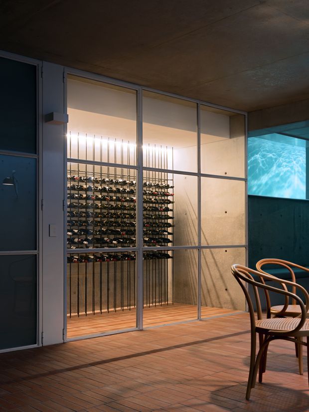 A dimly lit, contemporary wine room viewed through full-height framed glazing. Multiple horizontal bottle racks are mounted against a warm concrete or plaster wall, with vertical metal supports and timber flooring inside. The surrounding space has terracotta-toned tiled flooring, dark concrete surfaces, bentwood café chairs, and a blue-lit water feature or pool visible at right. The atmosphere is moody, refined, and highly architectural.