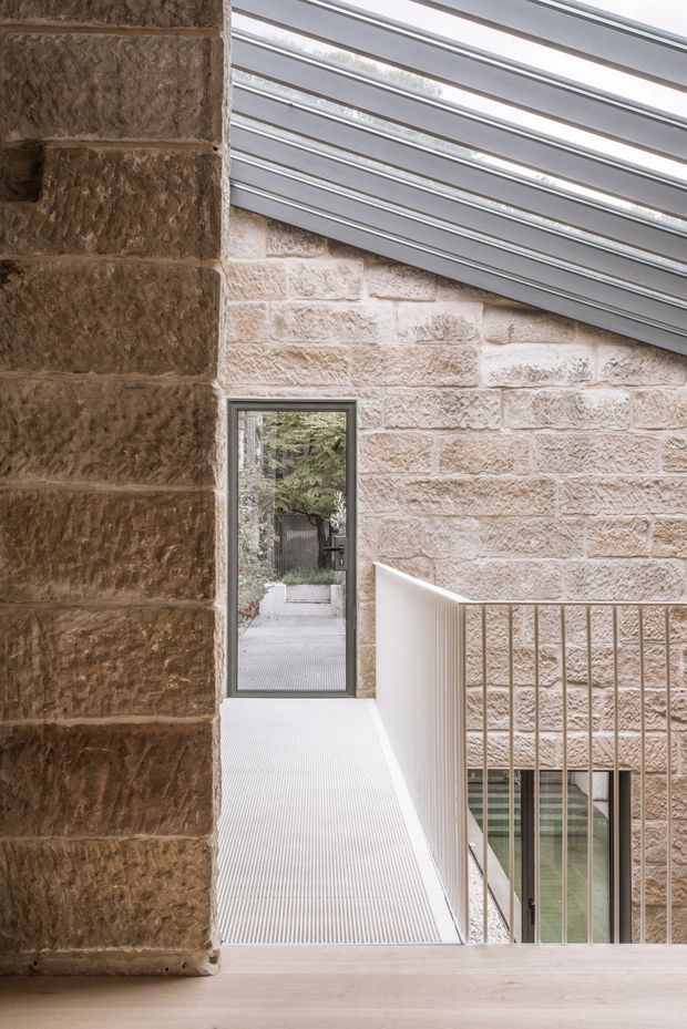 A narrow interior landing or walkway beside exposed sandstone masonry walls. The space features a sloped translucent roof with grey metal framing, a slim glazed door leading outdoors, a light ribbed floor surface, and a white balustrade overlooking a lower level. The image highlights an adaptive architectural connection between historic masonry and contemporary glazing and metalwork.