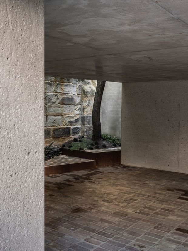 A sheltered architectural courtyard or covered outdoor passage with raw concrete walls and ceiling, a dark brick-paved floor, and a raised planted bed containing a tree. An exposed, weathered stone wall forms the backdrop, creating a restrained, monolithic composition with strong material contrast between concrete, brick, and stone.