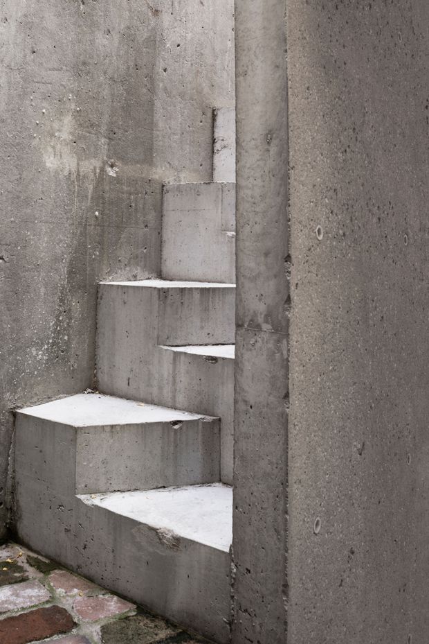 A narrow sculptural concrete stairway tucked between tall raw concrete walls, with irregular wedge-shaped steps rising from a small brick-paved area. The exposed concrete has a rough, unfinished texture and muted grey tone, creating a stark, minimalist architectural composition.