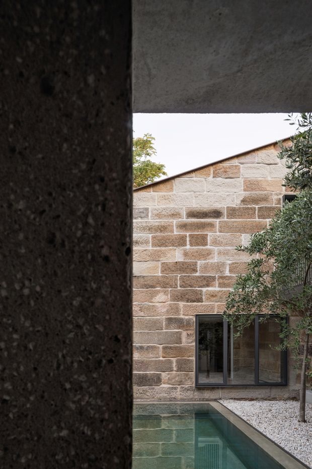 A narrow architectural view into a secluded courtyard framed by dark textured concrete walls and an overhead concrete slab. The courtyard features a small rectangular reflecting or swimming pool, pale gravel, a mature olive-like tree, and a sandstone block facade with a low sloping roof and dark-framed window. The composition is quiet and highly material-focused, with strong contrast between the dark concrete foreground and warm masonry beyond.