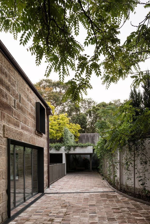 A narrow landscaped courtyard and paved driveway beside a stone-clad building. The scene features warm textured masonry walls, tall black-framed glazing, a brick-paved ground plane with linear drainage grates, planted boundary walls, mature overhanging trees, and a dark recessed covered entry or garage-like opening at the end of the passage. The image has a quiet, enclosed garden-courtyard character with contemporary detailing.