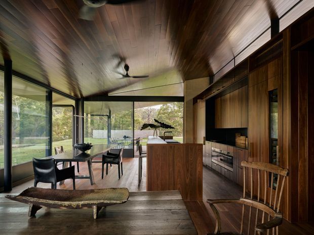Open-plan contemporary interior combining a kitchen, dining area and living space. Full-height glazing opens the room to a leafy outdoor setting, while extensive dark timber wall and ceiling panelling, built-in cabinetry, a central kitchen island and ceiling fans create a warm, natural atmosphere. The room includes a dining table, sculptural live-edge coffee table and timber lounge chair.