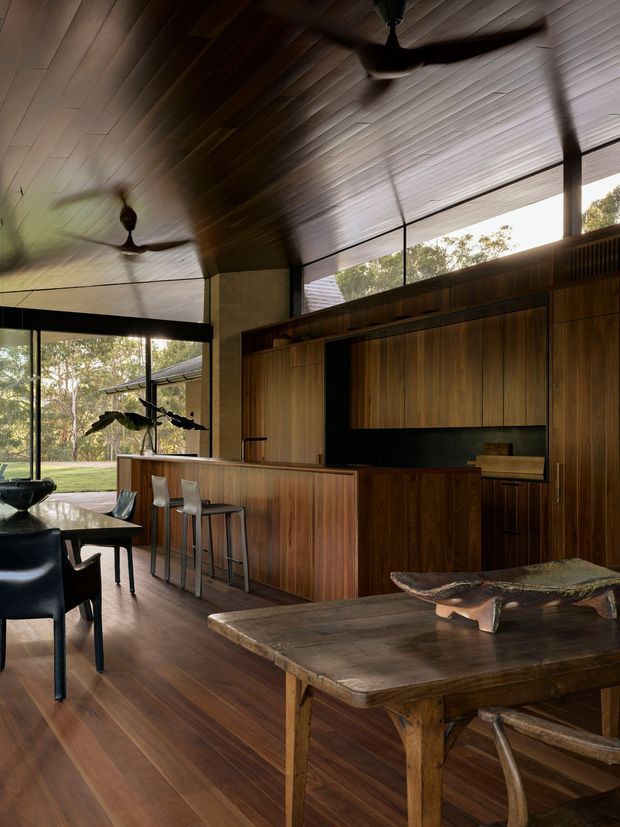 A warm, open-plan kitchen and dining area with extensive dark timber cabinetry, a long island with bar seating, timber flooring, and a high angled timber-lined ceiling. Large glazed openings connect the interior to a leafy outdoor setting. Ceiling fans, black-framed glazing, sculptural furniture, and a rustic dining table create a refined yet organic atmosphere.