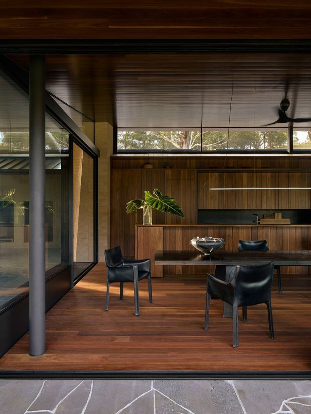 A warm, dark-toned open-plan dining and kitchen space with extensive timber lining and cabinetry. A dining table with black leather chairs sits in the foreground, while a long built-in kitchen and bar joinery occupies the rear wall. Full-height black-framed glazing opens toward a landscaped setting, and a dark timber ceiling with an integrated ceiling fan reinforces the strong indoor-outdoor connection. The space has a refined mid-century-influenced character with exposed structural posts and broad horizontal clerestory glazing.