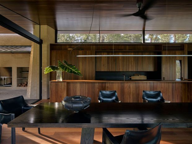A sophisticated open-plan dining and entertaining area with a large dark timber dining table, black leather dining chairs, and a long built-in timber bar or kitchen joinery wall. Rich vertical timber cabinetry, a timber-lined ceiling, expansive clerestory glazing, and dark structural framing create a warm, refined mid-century-inspired interior. A fireplace or outdoor cooking area is visible through the opening at left, while a ceiling fan and indoor greenery add functional and organic details.