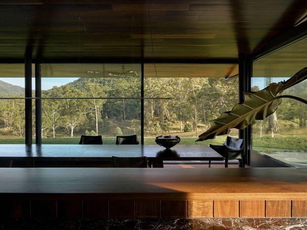 A dark, refined open-plan interior centered on a long timber dining or kitchen island with vertical timber detailing and a dark stone base. Full-height glazing frames a leafy bushland and water outlook, while a timber-lined ceiling, slim dark window frames, dining chairs, a sculptural bowl, and a large sculptural leaf form create a dramatic connection to the landscape.