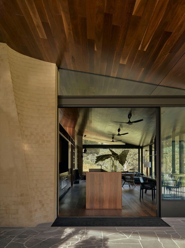View into a refined open-plan residential interior through a large dark-framed glazed opening. The space features a richly stained timber ceiling and floor, a curved pale brick or masonry wall, dark built-in cabinetry, a central timber island or counter, dining and lounge furniture, ceiling fans, and extensive glazing looking onto greenery. The image is architectural and warm, with strong indoor-outdoor connection and layered timber and masonry finishes.