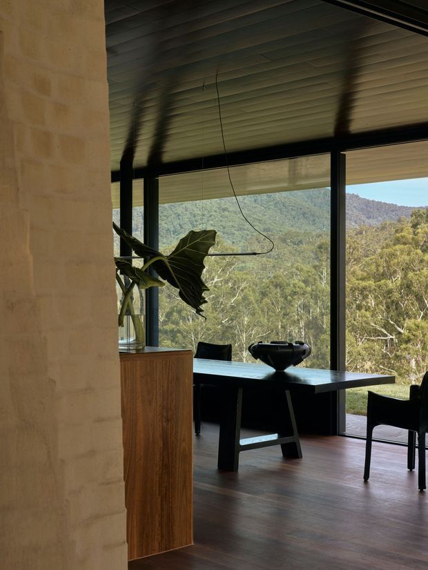 A moody contemporary dining area set against expansive floor-to-ceiling glazing overlooking a wooded, mountainous landscape. The room features a dark timber dining table with black chairs, a warm timber sideboard, a large sculptural plant, a pale masonry wall, dark framed windows, and a ribbed dark ceiling with concealed or minimal lighting.
