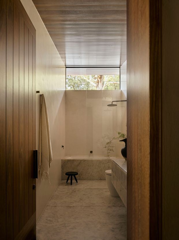 A narrow, warmly lit contemporary bathroom viewed through a timber doorway. The room features extensive pale natural stone or marble-like wall and floor surfaces, a built-in bath at the rear, a wall-mounted shower, toilet and vanity, vertical timber wall panelling, and a ribbed timber ceiling. A high horizontal window brings in filtered views of trees, while a towel hangs on the paneled wall and a small dark stool sits near the bath.